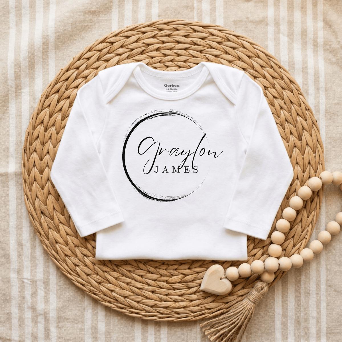 Introducing the Toodleroos Personalized Name Onesie – Custom Baby Bodysuit, shown here in white with “Graylon James” featured in elegant black script inside a bold black circle. The onesie is displayed on a woven placemat accented with wooden beads and a heart tassel, all set against a striped beige cloth background. This custom baby bodysuit makes the perfect announcement onesie to celebrate your little one’s special moments.
