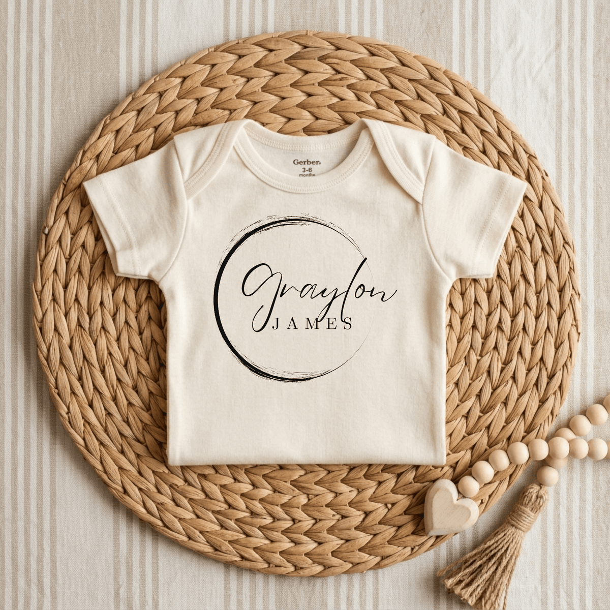 The Toodleroos Personalized Name Onesie – Custom Baby Bodysuit features “Graylon James” in elegant script inside a black circle on a cream baby bodysuit, beautifully displayed on a woven placemat with a wooden bead garland beside it—a charming custom piece perfect for announcing your little one’s arrival.