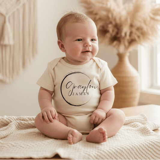 A baby wearing the Toodleroos Personalized Name Onesie – Custom Baby Bodysuit in cream sits on a knitted blanket. The onesie features the name Graylon James in script inside a circle. Blurred pampas grass and a vase decorate the background.