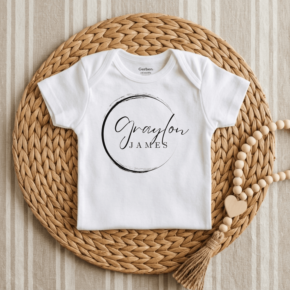 A Personalized Name Onesie – Custom Baby Bodysuit by Toodleroos, featuring the name Graylon James in a circular design, is displayed centered on a woven rattan placemat over a striped fabric surface, with a strand of wooden beads and a heart charm placed to the side.