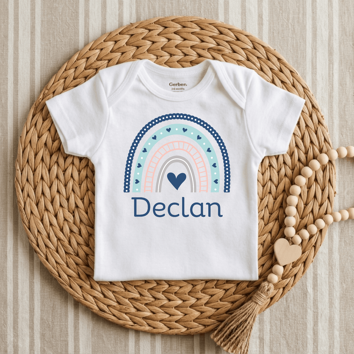 The Toodleroos Personalized Rainbow Baby Onesie – Custom Name Bodysuit, customized with the name Declan, features a charming boho rainbow and heart design. It is displayed on a woven placemat beside a beaded tassel decoration atop a striped fabric background—making it the perfect custom baby shower gift.