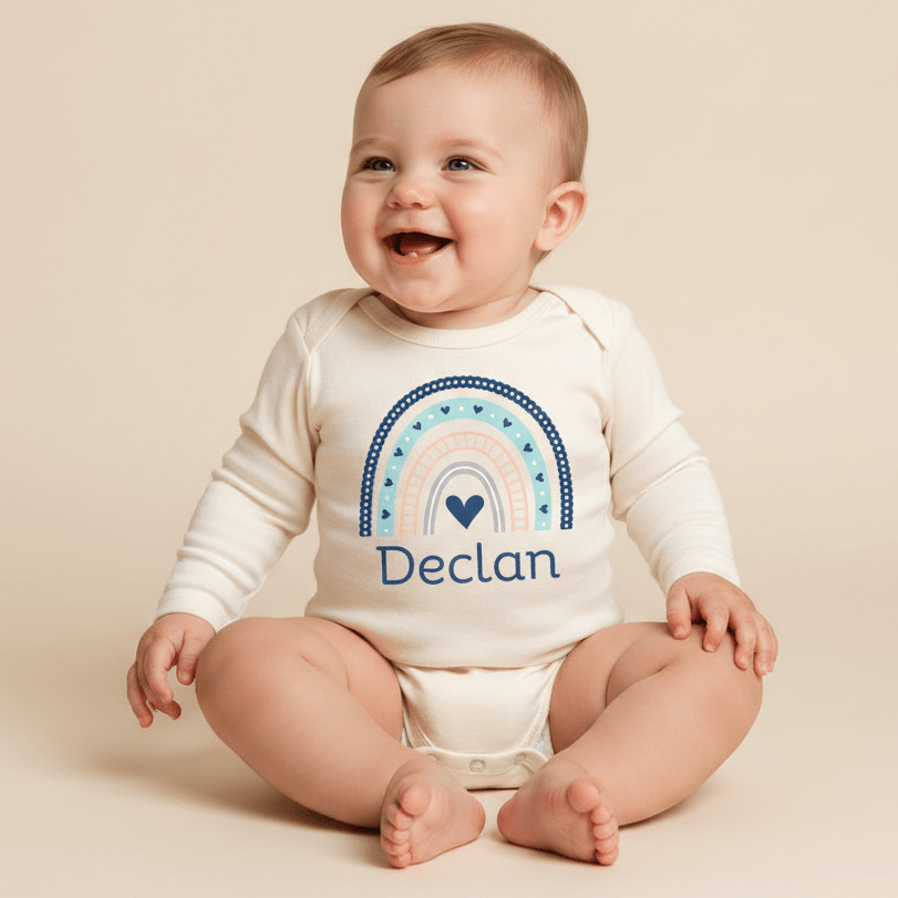 A smiling baby sits on a light beige background wearing the Toodleroos Personalized Rainbow Baby Onesie – Custom Name Bodysuit in a soft cream color, featuring a boho rainbow graphic and the name “Declan” printed on the front. This adorable outfit makes a perfect custom baby shower gift.