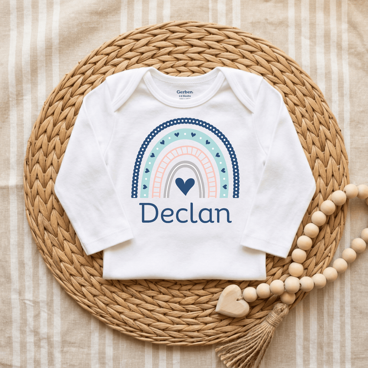 A Toodleroos Personalized Rainbow Baby Onesie – Custom Name Bodysuit, showcasing a boho rainbow and heart design with the name Declan printed on it, is displayed on a woven mat with a wooden beaded garland beside it. The striped fabric background highlights this charming and unique custom baby shower gift.