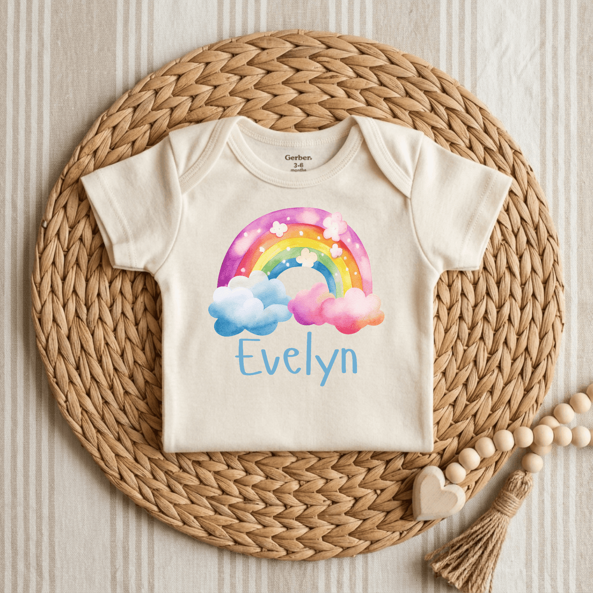 A cream-colored Personalized Rainbow Cloud Baby Girl Onesie by Toodleroos features a charming watercolor rainbow and clouds design. The custom name “Evelyn” is printed in blue beneath the rainbow, making this Custom Name Bodysuit a meaningful baby shower gift. The onesie is displayed on a round woven placemat atop a striped fabric background, with a wooden bead tassel accessory placed nearby.