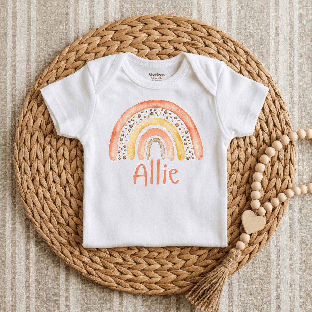 The Toodleroos Personalized Sunset Rainbow Newborn Onesie – Custom Name Bodysuit features a pastel rainbow and the name Allie printed on soft white fabric, displayed on a woven mat with wooden beads and a heart—making it the ideal baby shower gift or coming-home outfit.