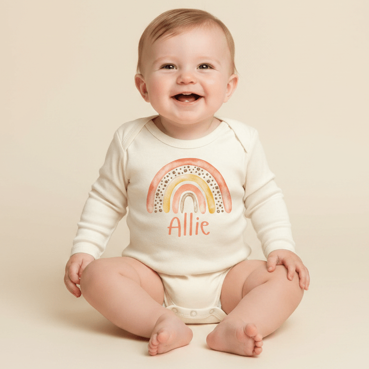 A smiling baby sits on a cream background wearing the Toodleroos Personalized Sunset Rainbow Newborn Onesie – Custom Name Bodysuit, featuring a vibrant rainbow design and “Allie” printed across the front—perfect for a memorable baby shower gift or an adorable coming-home outfit.