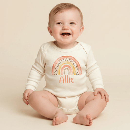 A smiling baby sits on a cream background wearing the Toodleroos Personalized Sunset Rainbow Newborn Onesie – Custom Name Bodysuit, featuring a vibrant rainbow design and “Allie” printed across the front—perfect for a memorable baby shower gift or an adorable coming-home outfit.