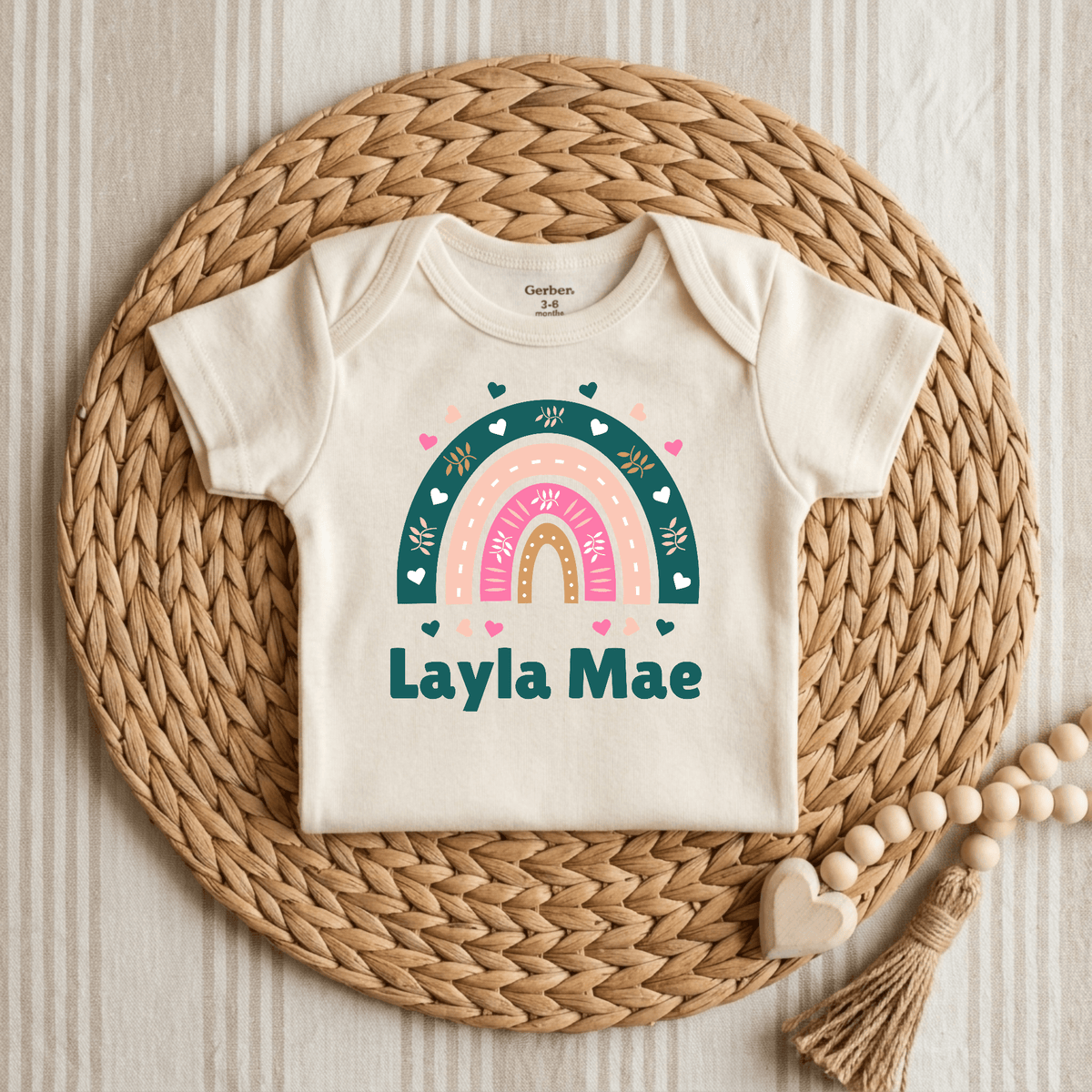 An off-white short-sleeve baby bodysuit laid flat on a round woven straw mat. The bodysuit has a colorful rainbow graphic with hearts and the text 'Layla Mae' in teal.