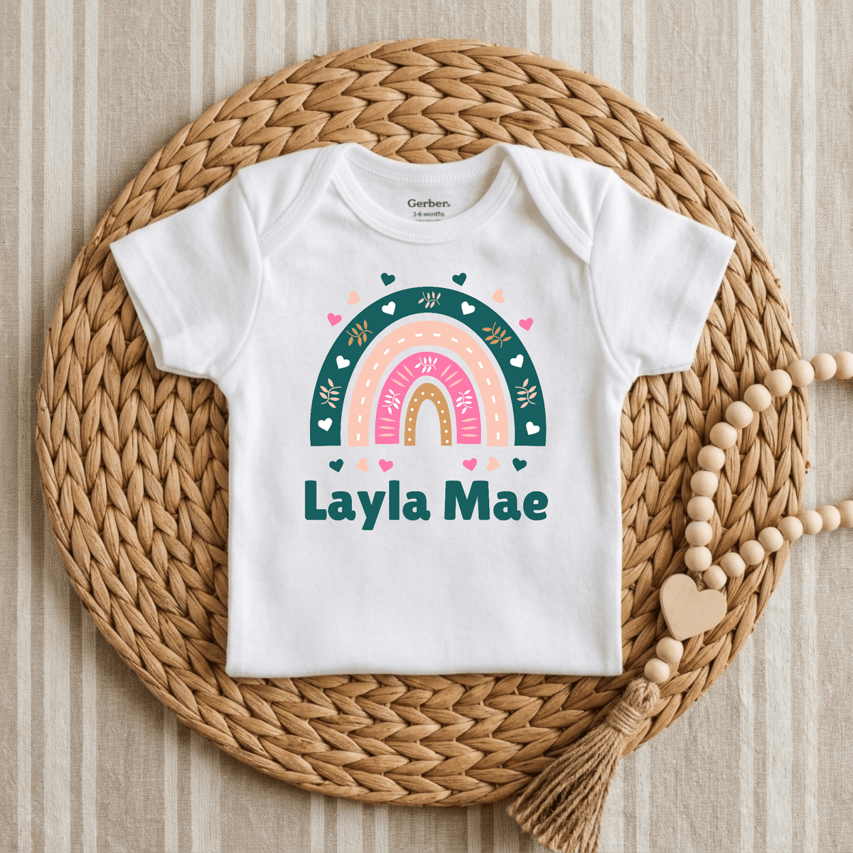 A white Gerber short-sleeve baby bodysuit with a colorful rainbow graphic and the text 'Layla Mae' printed on the front, styled flat on a round woven straw mat with a wooden bead garland.