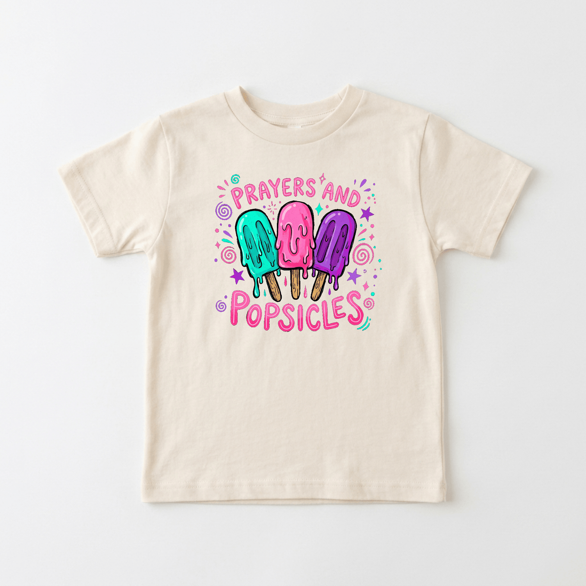 The Toodleroos Prayers and Popsicles Kids Shirt – Summer Faith Tee features a beige t-shirt adorned with colorful cartoon popsicles, pink “Prayers and Popsicles” text, and charming decorative accents on a plain white background—an ideal faith-themed kids shirt or cheerful choice for Vacation Bible School.