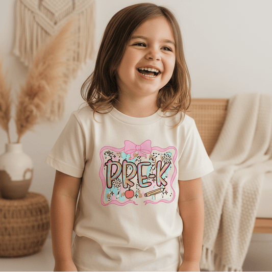 A smiling young girl with brown hair wears the Toodleroos Pre-K Youth Tee – Bow & Apple Back to School Shirt, a colorful and fun choice for a first day of school outfit, as she stands in a cozy room decorated with soft neutrals and woven accents.