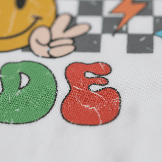 Capture the playful spirit of the Toodleroos Preschool Dude Kids Shirt – Back to School Tee with a close-up of colorful, textured “DE” letters on fabric, featuring design elements like a yellow smiley face, peace sign hand, checkerboard pattern, and an orange lightning bolt in the background—perfect for a fun and retro kids tee.