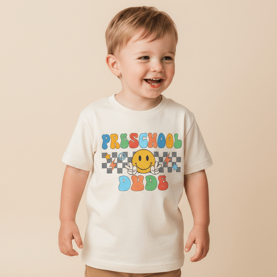 A smiling young child stands against a beige background, wearing the Toodleroos Preschool Dude Kids Shirt – Back to School Tee—a white shirt featuring colorful retro text that reads “Preschool Dude” and a playful yellow smiley face with wings.