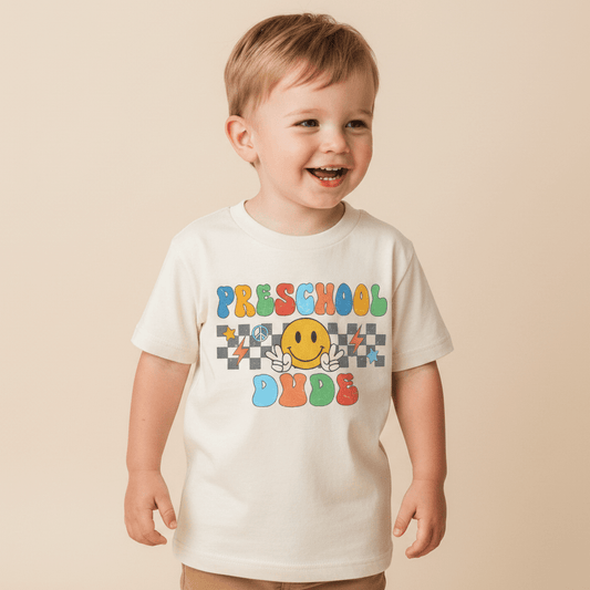 A smiling young child stands against a beige background, wearing the Toodleroos Preschool Dude Kids Shirt – Back to School Tee—a white shirt featuring colorful retro text that reads “Preschool Dude” and a playful yellow smiley face with wings.
