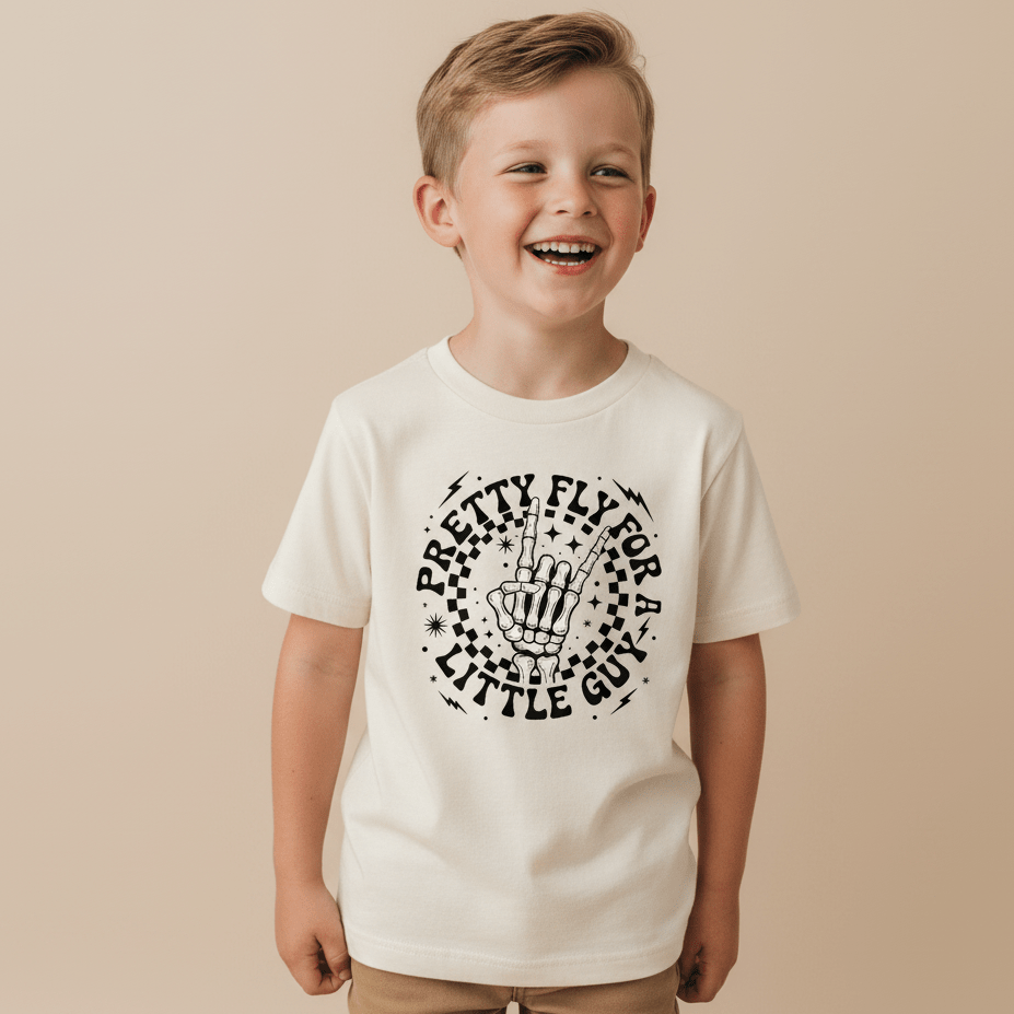 A smiling young boy wears the Toodleroos Pretty Fly for a Little Guy Kids Shirt – a white tee featuring a rock skeleton hand graphic and playful lettering – while standing against a plain beige background.