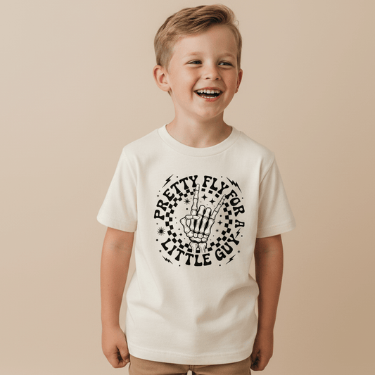 A smiling young boy wears the Toodleroos Pretty Fly for a Little Guy Kids Shirt – a white tee featuring a rock skeleton hand graphic and playful lettering – while standing against a plain beige background.