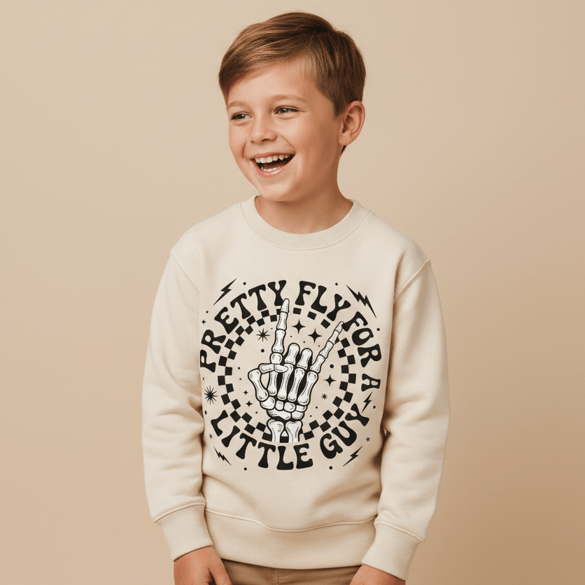 A smiling young boy wears the Toodleroos Pretty Fly for a Little Guy Kids Sweatshirt – Rock Skeleton Pullover, featuring a skeleton hand graphic and text on the front, while standing against a beige background.