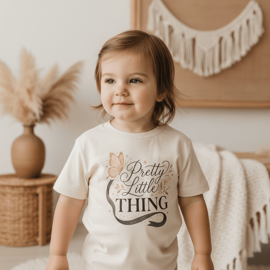 A toddler with brown hair wears the Toodleroos Pretty Little Thing Toddler Tee – Cute Butterfly Girls Shirt while standing indoors surrounded by neutral decor, including pampas grass and woven baskets in the background.