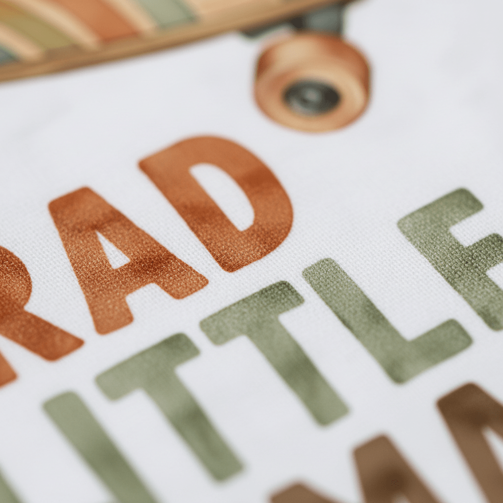 Close-up of vibrant text reading “RAD LITTLE HUMAN” on white fabric, featuring a partial skateboard graphic at the top. Bold orange and green lettering give the Toodleroos Toddler Skateboard Shirt – Rad Little Human Short-Sleeve Graphic Tee a playful retro look, making it the ideal toddler graphic tee for little skaters in training.