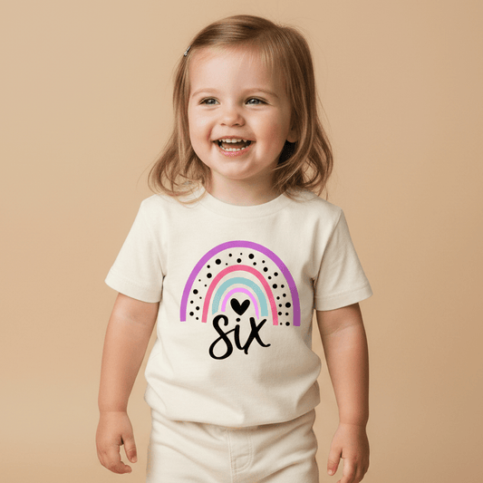 A smiling young child dressed in cream stands before a tan backdrop, wearing the Toodleroos Rainbow 6th Birthday Kids Shirt—a cheerful 6th birthday tee featuring a pink rainbow, black dots, a heart, and “six” written below the rainbow.