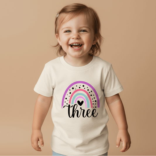 A smiling toddler with light brown hair wears the Toodleroos Rainbow Three Toddler Shirt – 3rd Birthday Tee, featuring a pink and green rainbow and the word “three” on a cream background, standing against a beige backdrop.