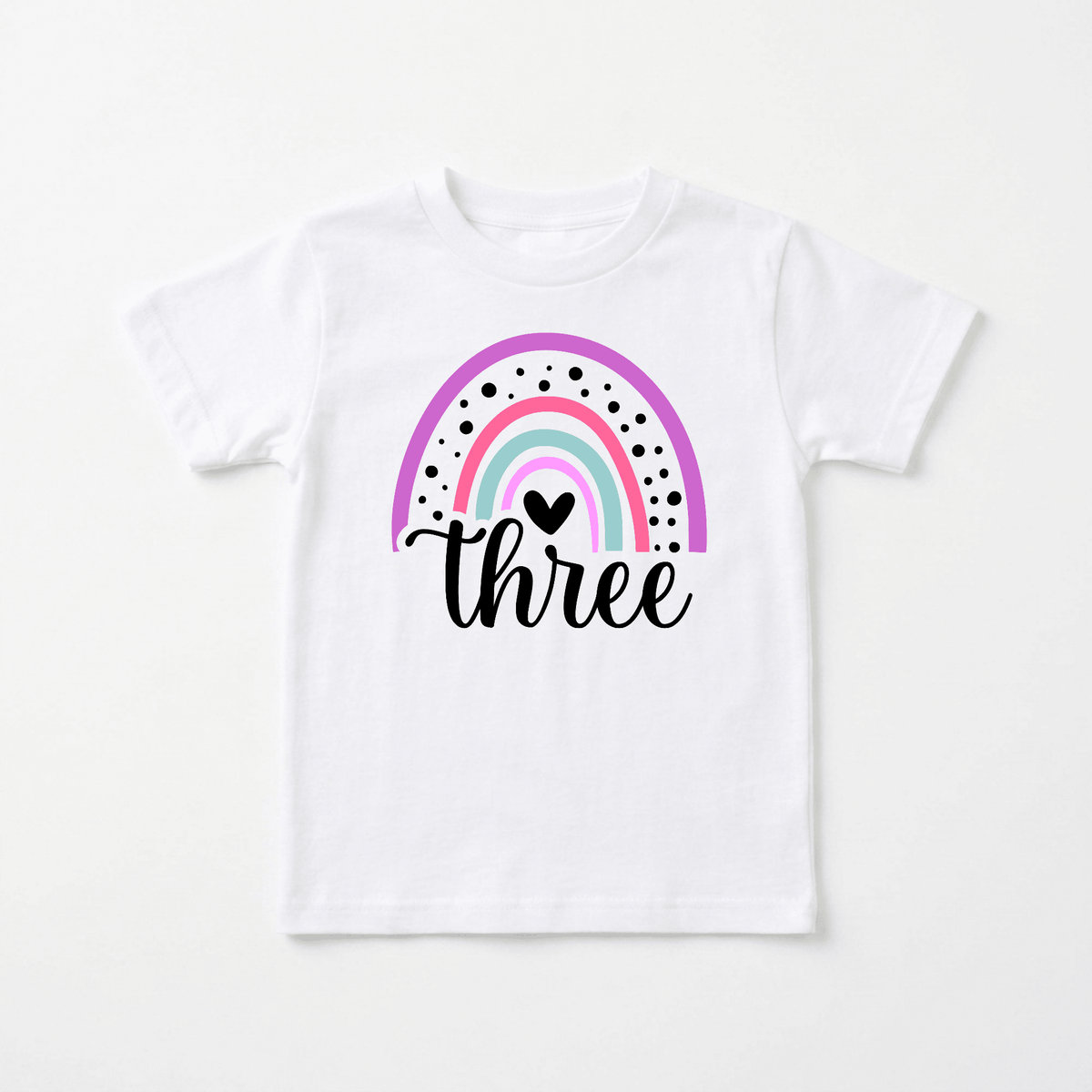 Introducing the Rainbow Three Toddler Shirt – 3rd Birthday Tee by Toodleroos: a white tee showcasing a pastel rainbow, black dots, a black heart, and the word “three” in a playful script beneath the rainbow. This delightful shirt is perfect as a 3rd birthday outfit or for anyone seeking personalized kids’ shirts with whimsical charm.