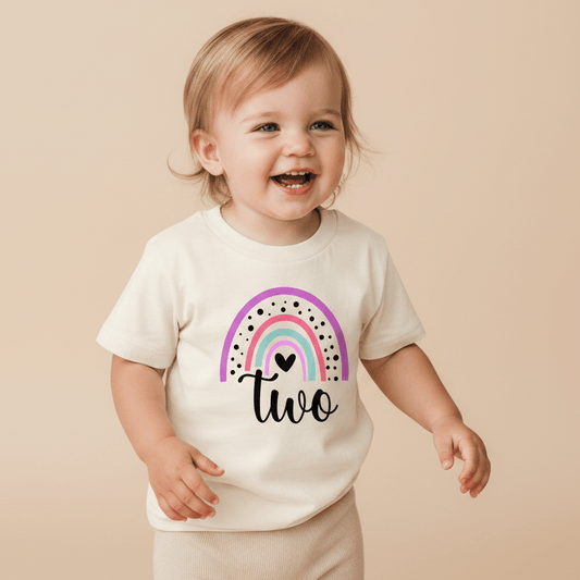 A smiling toddler wears the Toodleroos Rainbow Two Toddler Shirt – 2nd Birthday Tee, featuring a vibrant design and the word “two” on a cream-colored background, standing against a beige backdrop. This cheerful shirt is perfect for celebrating your little one’s 2nd birthday milestone.