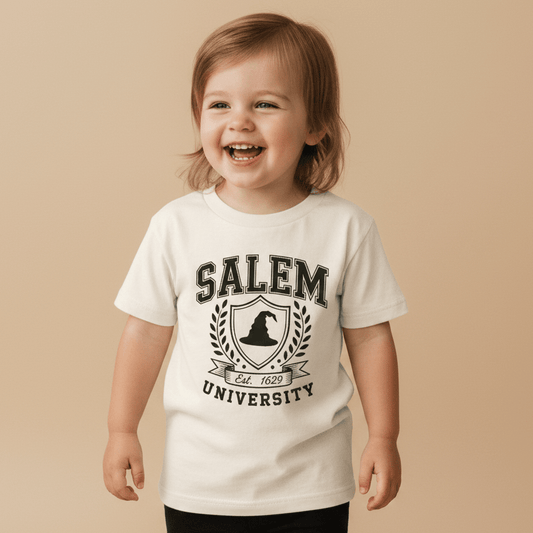 A cheerful toddler models the Toodleroos Salem University Toddler Shirt – Witch Hat Crest Tee, featuring a white design with a witch hat crest and laurel motif, posed against a beige background.