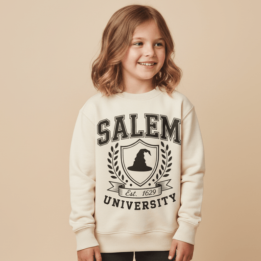 A smiling young girl with wavy hair wears the Toodleroos Salem University Toddler Sweatshirt in cream, featuring a witch hat crest and “Est. 1629” on the front, set against a neutral background.