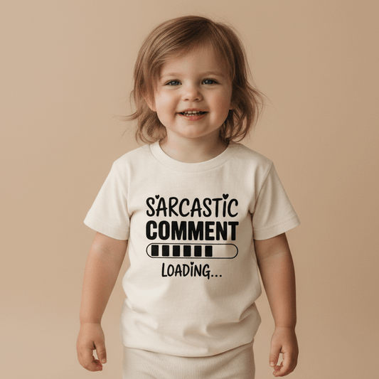 A smiling toddler wears the Toodleroos “Sarcastic Comment Loading Toddler Shirt – Funny Loading Bar Tee,” featuring the phrase “Sarcastic Comment Loading...” and a loading bar graphic, while standing against a plain beige background.