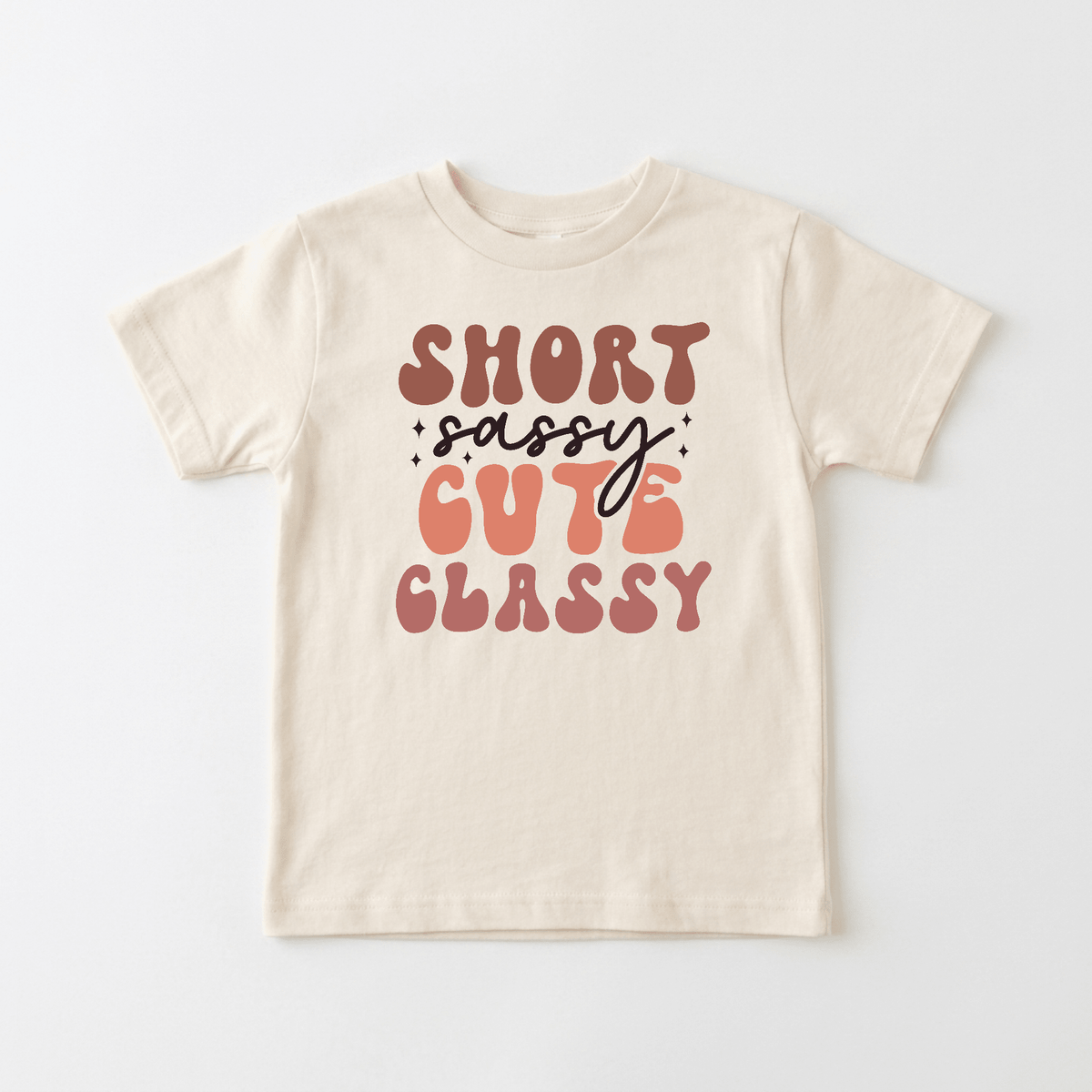 The Toodleroos Short Sassy Cute Classy Kids Shirt – Retro Tee features a beige background with bold, colorful text in playful fonts that reads “Short Sassy Cute Classy.” The vibrant pink and brown lettering is accented with decorative sparkles around “sassy,” giving this vintage-inspired kids tee a fun and lively vibe. Perfect for children who love retro shirts with extra personality.