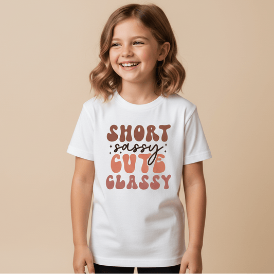 A cheerful young girl with light brown hair wears the Toodleroos Short Sassy Cute Classy Kids Shirt – Retro Tee, featuring playful, colorful retro lettering, standing against a neutral beige background for a fun, vintage-inspired kids tee vibe.