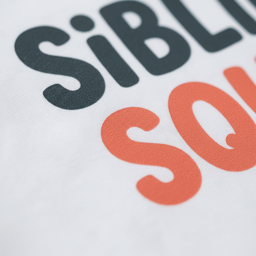 Close-up of text printed on white fabric, featuring the partial words SIBL in bold black and SO in vibrant orange. These modern letters suggest a larger message, making it perfect for the Toodleroos Sibling Squad Toddler Shirt – a playful matching sibling tee that’s ideal as a cozy toddler shirt.