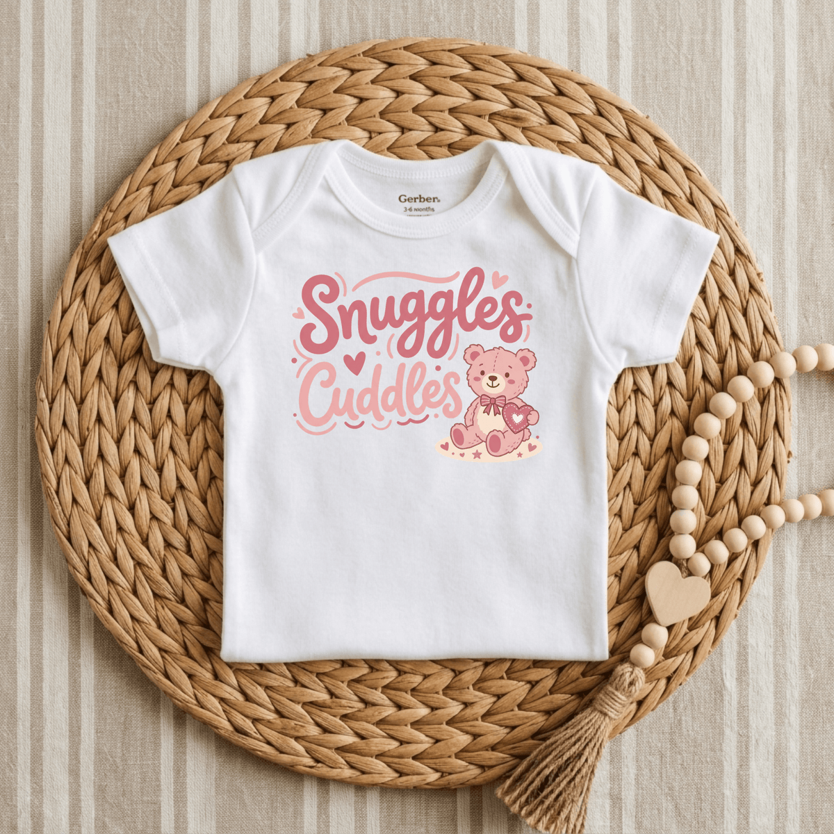The Toodleroos Snuggles & Cuddles Teddy Bear Onesie – Baby Girl Bodysuit features “Snuggles & Cuddles” in pink script with an adorable teddy bear, displayed on a woven placemat with a beaded tassel garland—an ideal baby shower gift or a sweet choice for anyone looking for cute baby onesies.