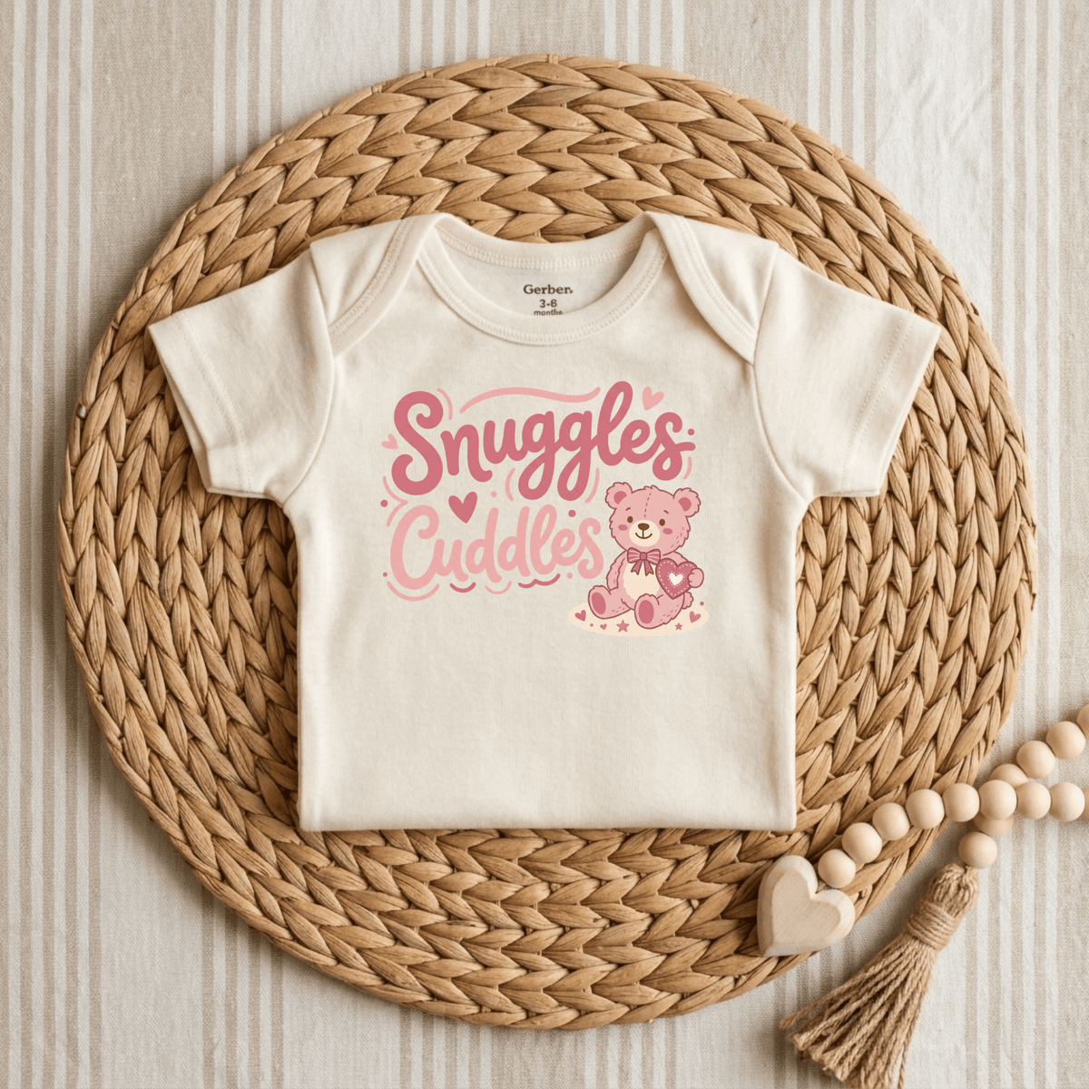 A cream-colored Snuggles & Cuddles Teddy Bear Onesie – Baby Girl Bodysuit by Toodleroos is displayed on a round woven placemat. This adorable onesie features pink “Snuggles Cuddles” text above a cartoon teddy bear holding a heart. Nearby, a wooden bead garland with a heart charm rests on striped fabric—making it the perfect baby shower gift.