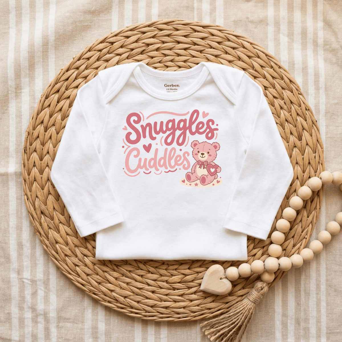 The Toodleroos Snuggles & Cuddles Teddy Bear Onesie – Baby Girl Bodysuit features charming pink “Snuggles & Cuddles” text and an adorable teddy bear on a white onesie, displayed on a woven mat with a wooden bead garland beside it atop a striped fabric background—making it the perfect baby shower gift or an irresistibly cute choice for anyone searching for adorable baby onesies.
