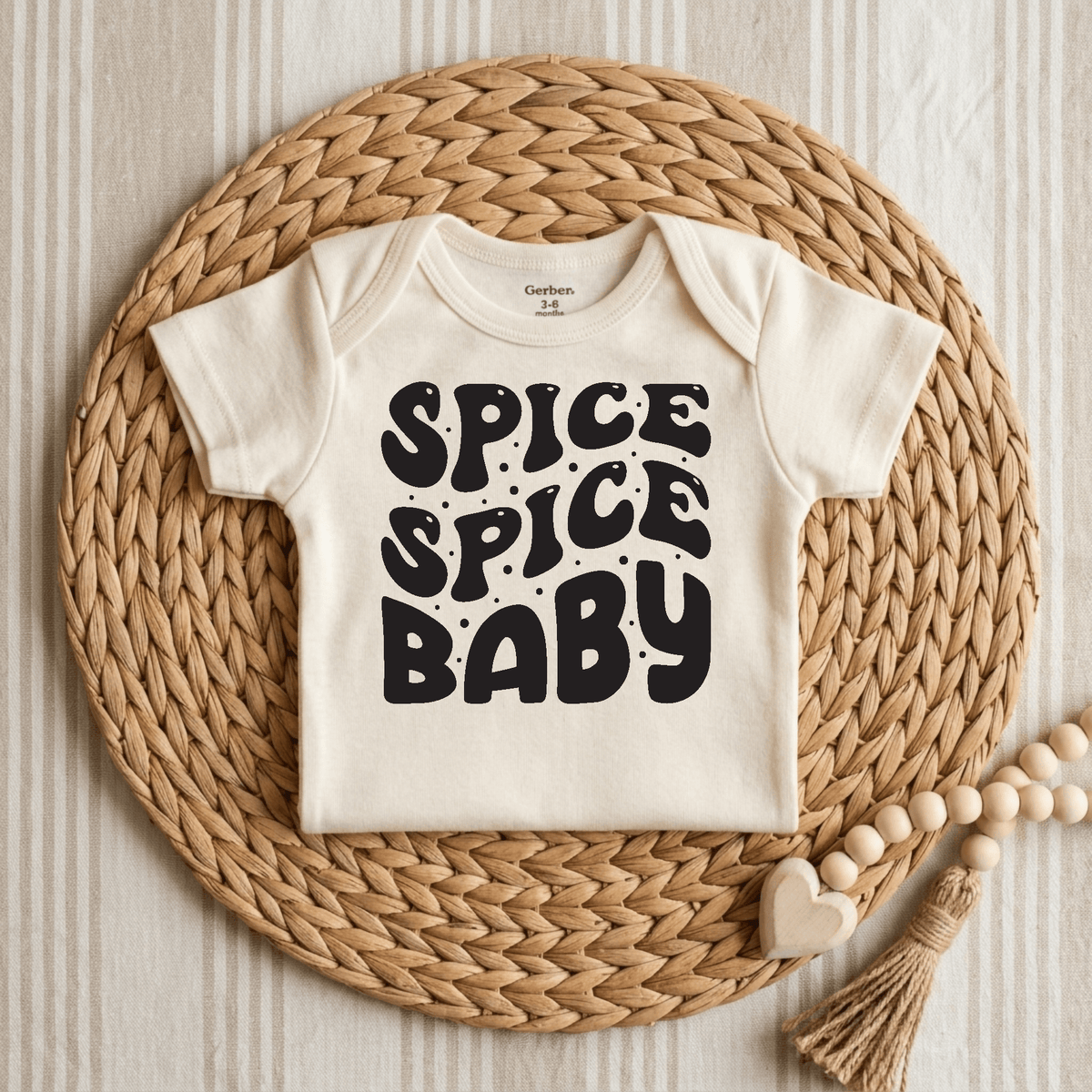 The Toodleroos Spice Spice Baby Onesie – Funny Retro Bodysuit, featuring bold black “Spice Spice Baby” text on a cream background, is styled atop a beige striped fabric with a woven placemat and wooden bead and heart garland. This gender-neutral outfit is perfect for adding playful retro flair to your baby’s wardrobe.