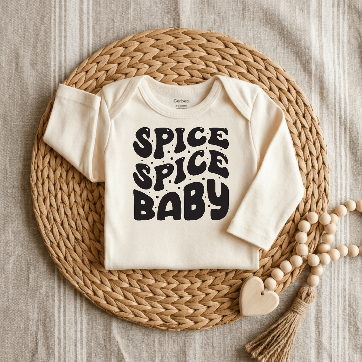 The Toodleroos Spice Spice Baby Onesie – Funny Retro Bodysuit features bold black “Spice Spice Baby” text on a cream-colored fabric, displayed on a woven placemat alongside a wooden bead and heart accessory. This gender-neutral baby outfit is perfect for little ones with big personality.