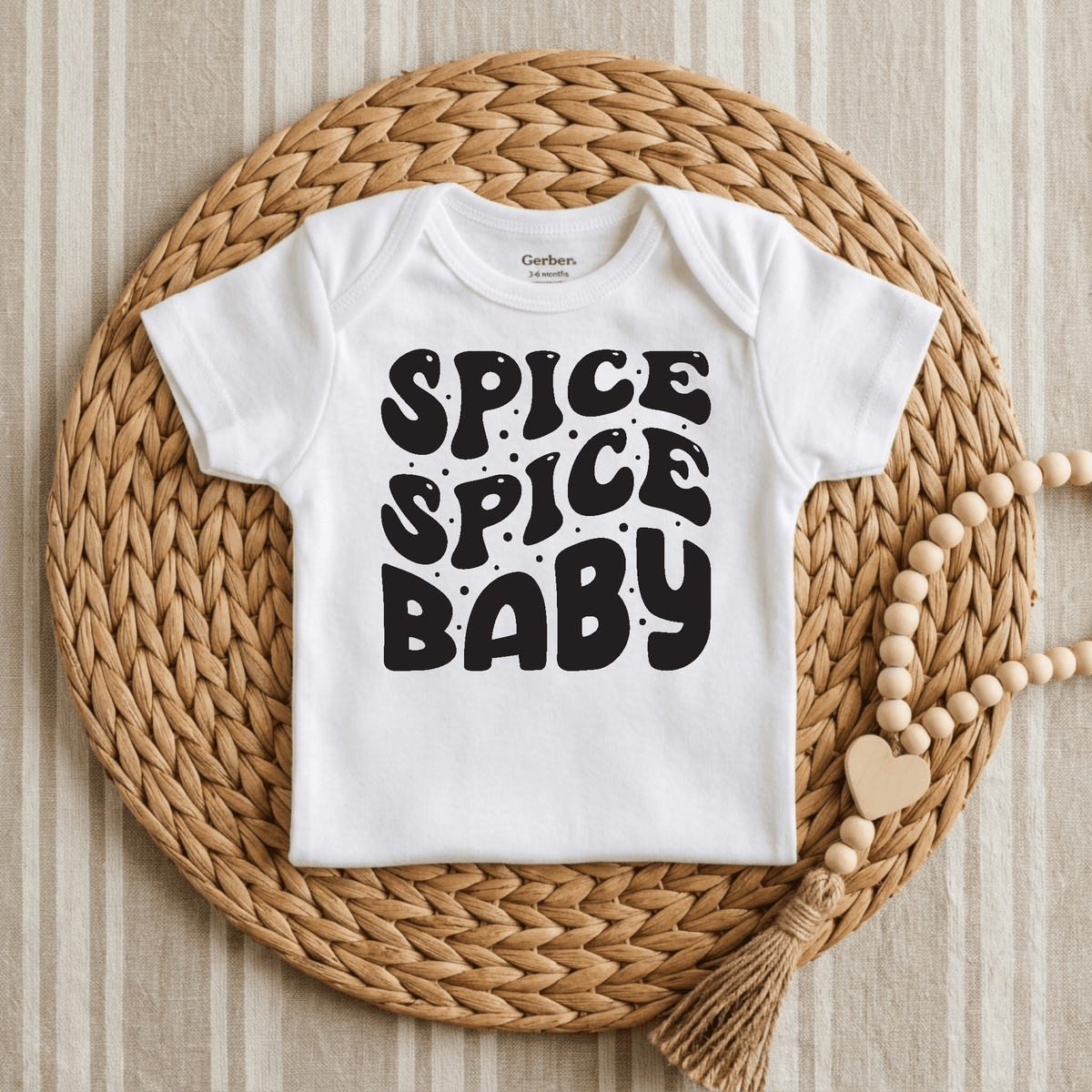 A white Toodleroos Spice Spice Baby Onesie – Funny Retro Bodysuit with bold black lettering spelling out “SPICE SPICE BABY” is displayed on a round woven placemat, accompanied by a beaded tassel accessory on a striped fabric background. This gender-neutral outfit from Toodleroos is an ideal choice for parents seeking playful and funny baby onesies to add some retro flair to their little one’s wardrobe.