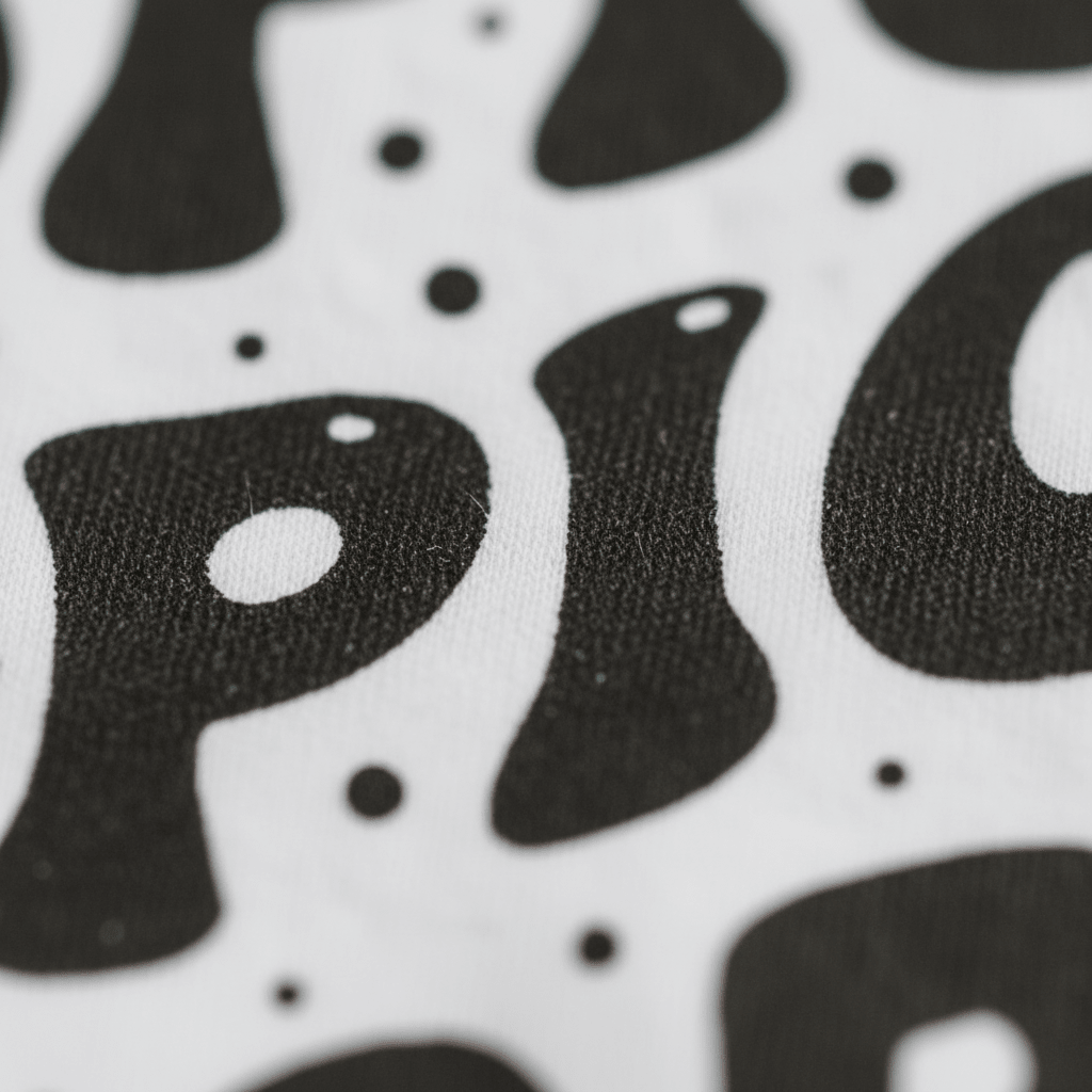 A close-up of black, blob-like letters and dots printed on white fabric, showing the textured surface and part of the word “Spice” in a playful font—perfect for the Toodleroos Spice Spice Baby Onesie, a funny retro bodysuit that’s ideal for gender-neutral baby outfits.