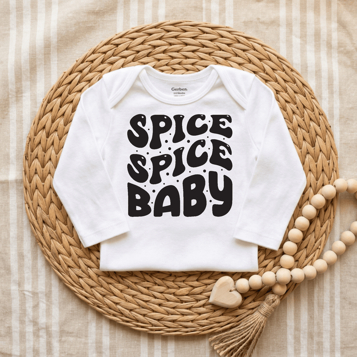 A white Toodleroos Spice Spice Baby Onesie – Funny Retro Bodysuit, featuring “Spice Spice Baby” in bold black letters, is displayed on a woven round mat with a tasseled wooden bead garland on a beige striped cloth. This gender-neutral bodysuit is one of the most amusing baby onesies, perfect for adding playful retro style to your little one’s wardrobe.