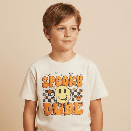A young boy stands against a plain beige background, wearing the Toodleroos Spooky Dude Retro Kids Shirt—a white Halloween tee featuring “Spooky Dude” in bold orange letters, a yellow melting smiley face, and festive graphics like pumpkins and bats.