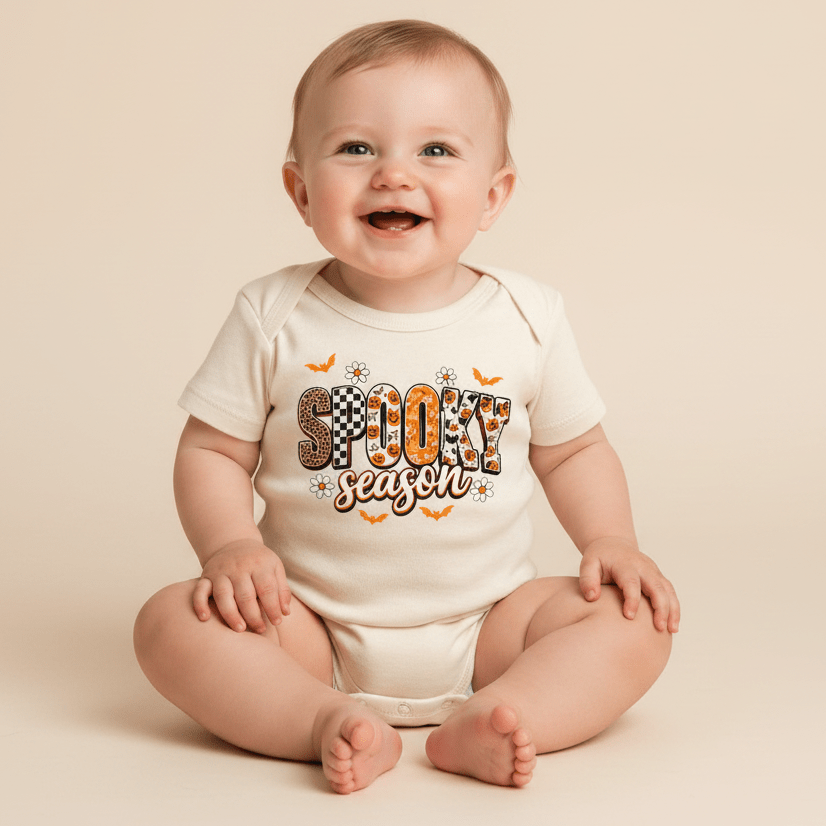 A happy baby sits on a beige background wearing the Toodleroos Spooky Season Baby Onesie – Trendy Halloween Bodysuit, featuring festive bat and white flower patterns—the ideal Halloween outfit for your little one to celebrate in style.
