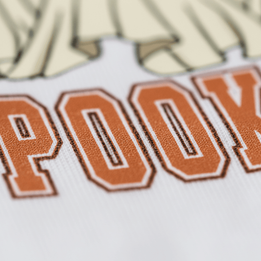 Close-up of orange varsity-style letters spelling POOK on soft white fabric, with cream-colored ghost shapes above the letters—part of the Toodleroos Spooky Vibes Dog Ghosts Kids Shirt, a fun Halloween tee perfect for kids.