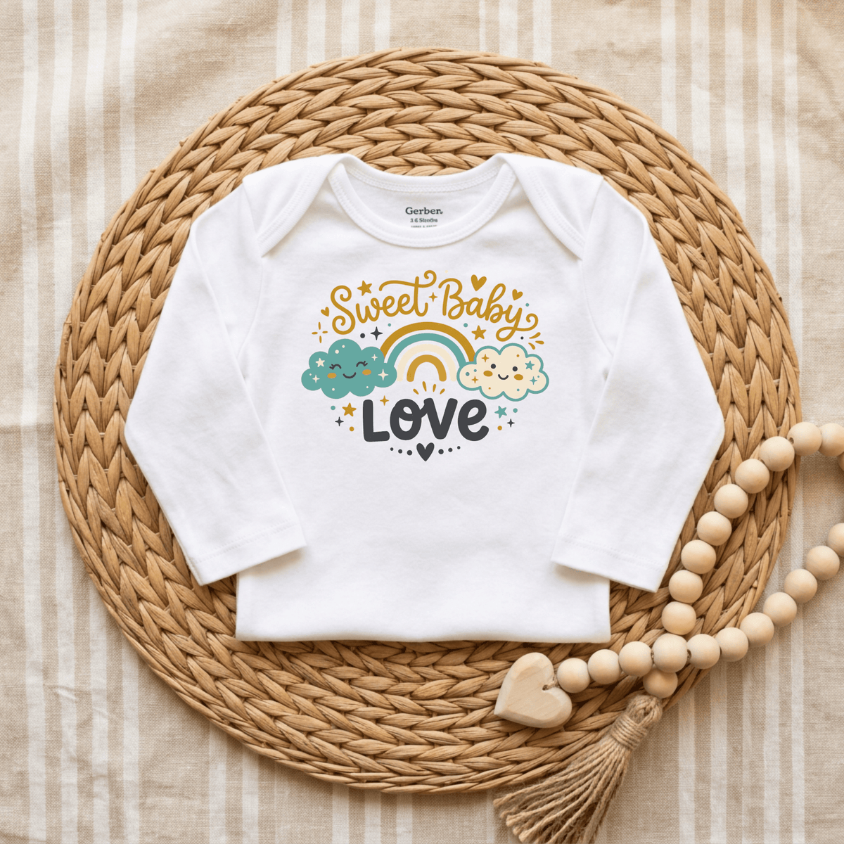 A Toodleroos Sweet Baby Love Onesie – Cute Rainbow Cloud Bodysuit, featuring a white, gender-neutral design with “Sweet Baby Love” and colorful clouds and rainbow accents, is displayed on a woven mat beside a wooden heart beaded decoration.