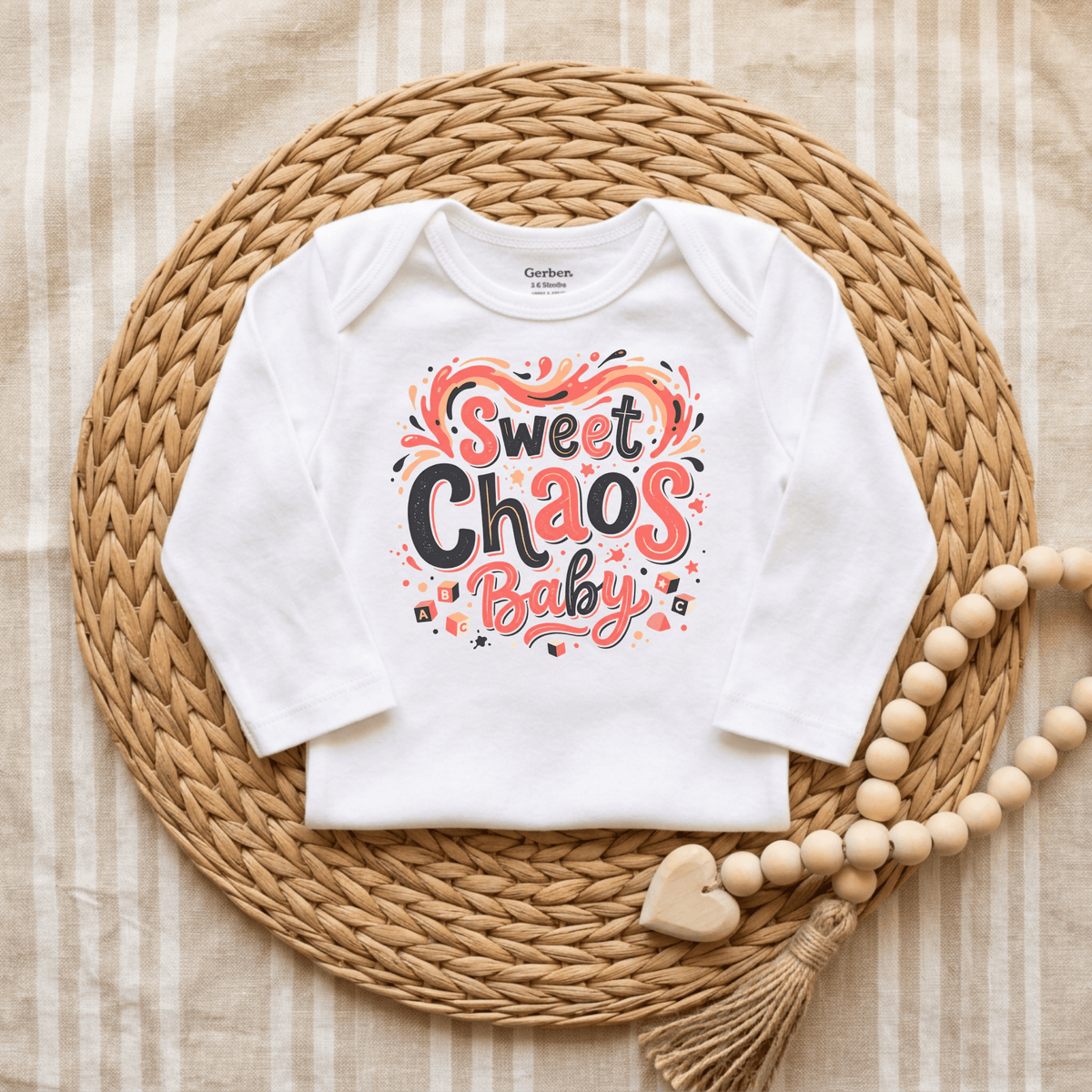 The Toodleroos Sweet Chaos Baby Onesie – Funny Long Sleeve Bodysuit features “Sweet Chaos Baby” in vibrant, playful letters on a crisp white fabric. Styled atop a woven mat with a wooden bead and heart tassel beside it, and set against a striped fabric background, this unisex onesie offers the perfect gender-neutral baby gift for anyone seeking funny baby onesies with adorable charm.