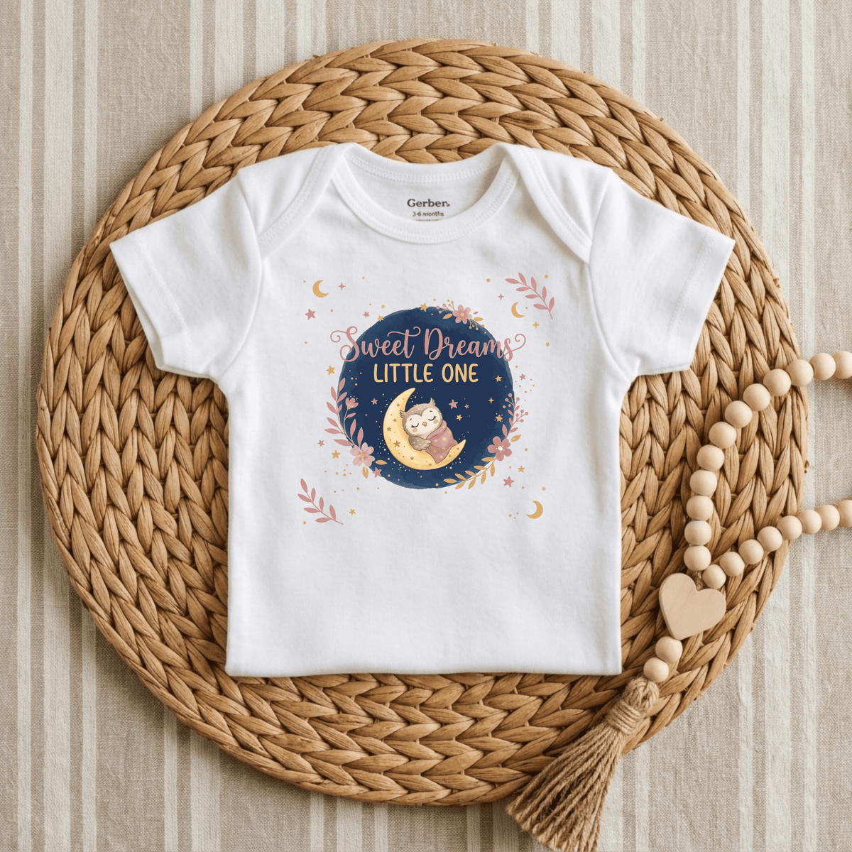 A white baby onesie from Toodleroos is displayed on a woven mat, adorned with a charming design of a sleeping owl and stars. This gender-neutral Sweet Dreams Little One Onesie – Cute Baby Owl Bodysuit features the phrase “Sweet Dreams Little One” and is accented by wooden beads and a heart shape beside it.