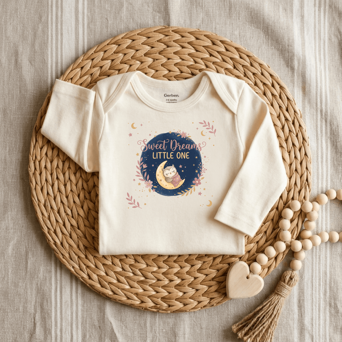 The Toodleroos Sweet Dreams Little One Onesie is a cream-colored, gender-neutral baby bodysuit featuring a cute owl design, shown on a woven mat alongside a wooden heart and bead garland.