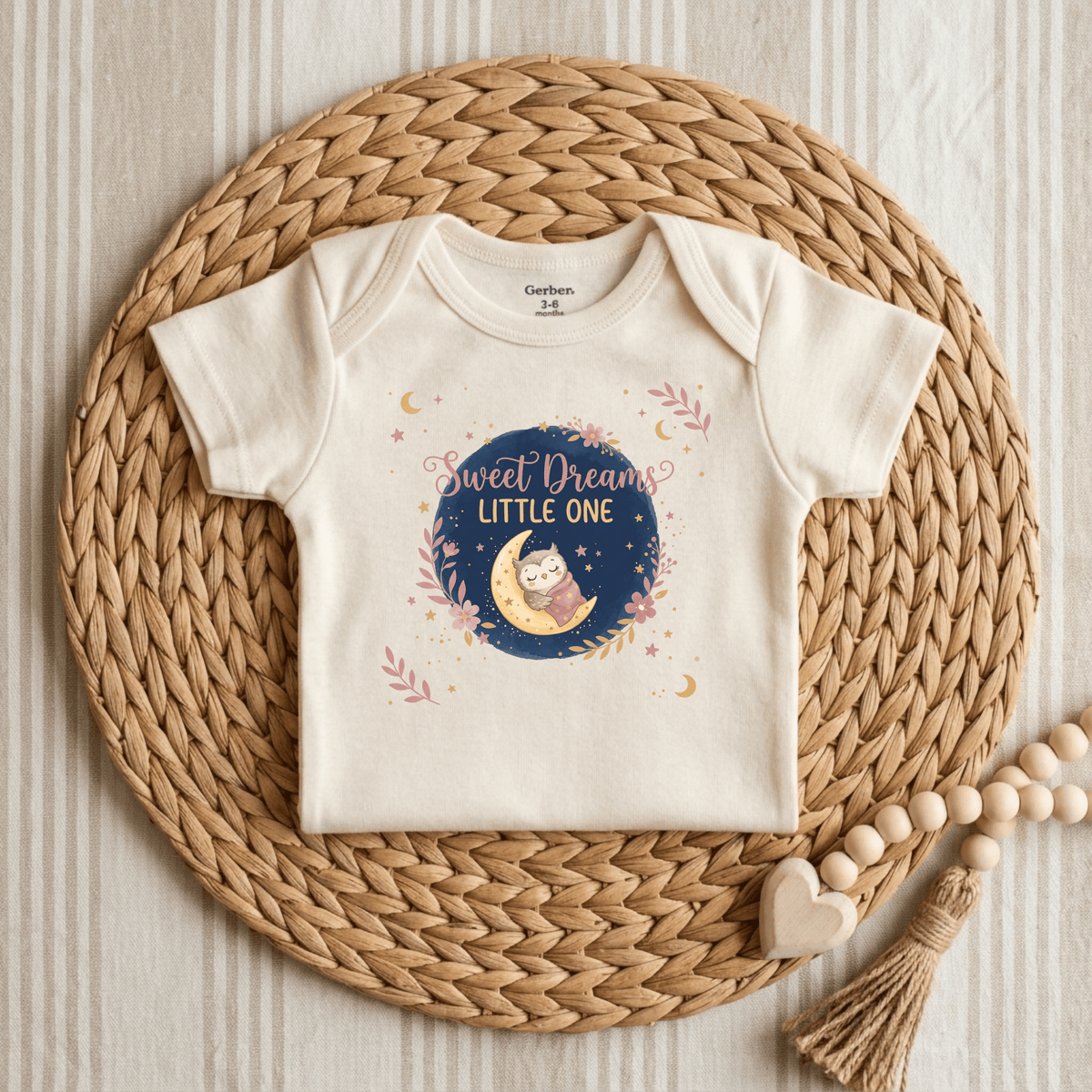 Introducing the Sweet Dreams Little One Onesie by Toodleroos – a cream-colored, gender-neutral baby bodysuit adorned with a sleepy owl nestled on a crescent moon, surrounded by scattered stars and the heartfelt phrase “Sweet Dreams Little One.” Shown here on a woven placemat next to a wooden teether, this adorable woodland-themed onesie is the perfect cozy addition to any nursery.
