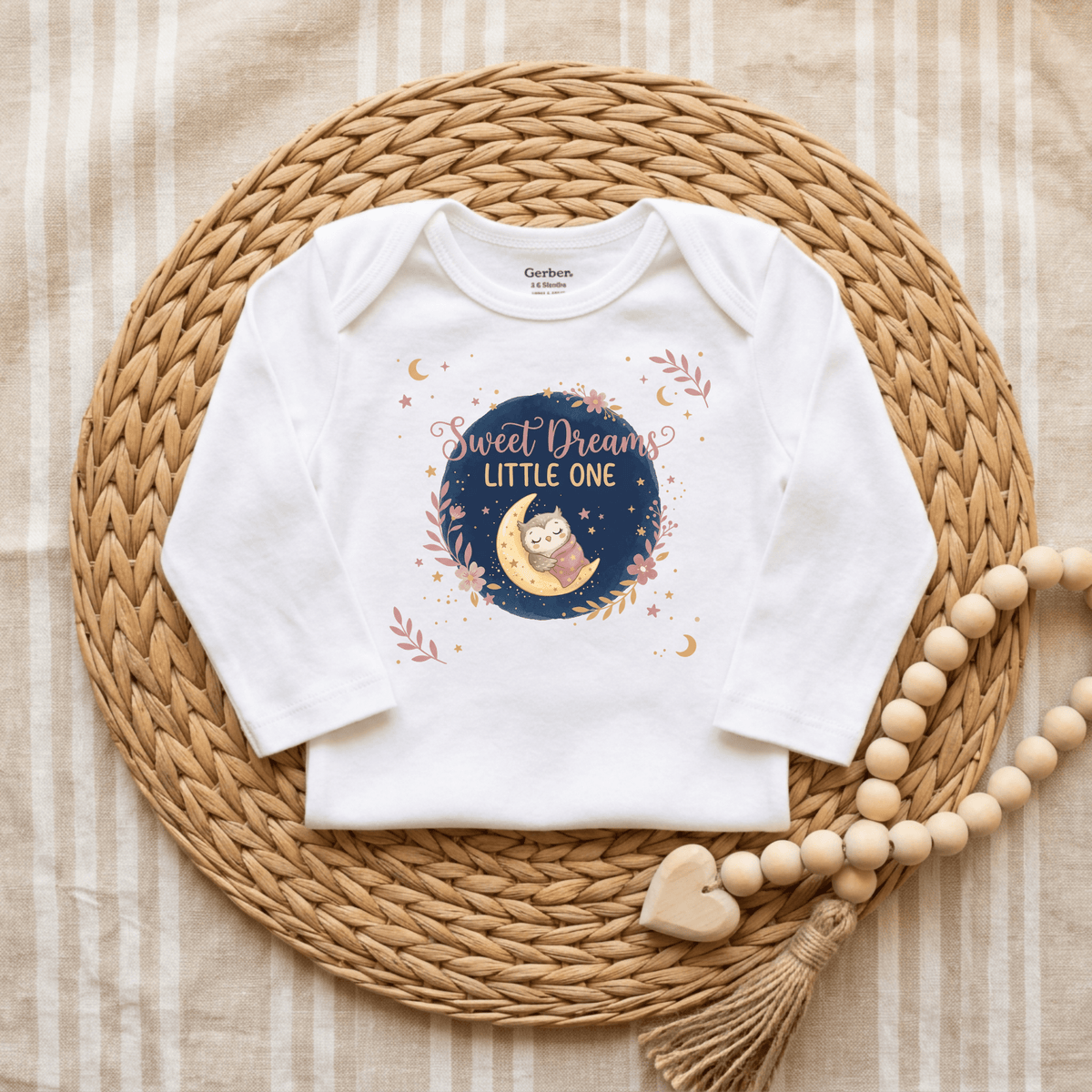 The Toodleroos “Sweet Dreams Little One” Onesie – a gender-neutral white baby bodysuit featuring an adorable baby owl design and cute moon and stars, is displayed on a woven mat beside a beaded tassel decoration.